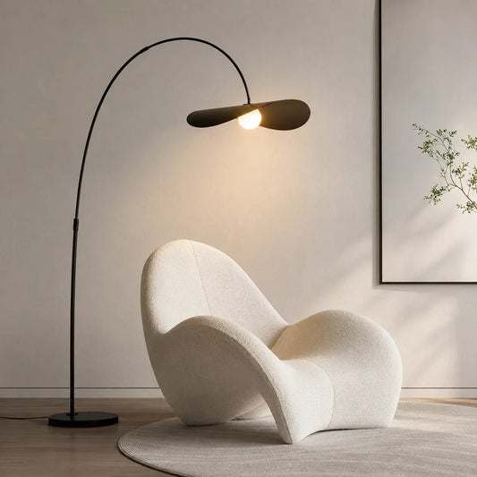 An adjustable sculptural arc floor lamp in black, casting a warm light over a modern cream bouclé armchair in a minimalist living room.