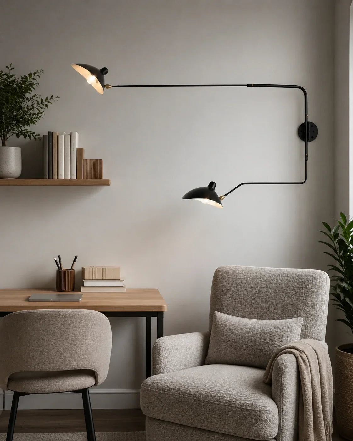 Black double swing-arm wall lamp with two adjustable shades, mounted on a wall above a grey reading armchair and wooden desk in a modern room.