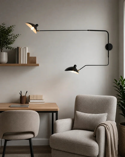 Black double swing-arm wall lamp with two adjustable shades, mounted on a wall above a grey reading armchair and wooden desk in a modern room.