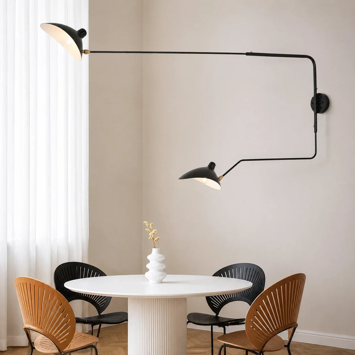 A modern black double swing-arm wall lamp mounted on a neutral wall above a round dining table with chairs in a contemporary dining area.