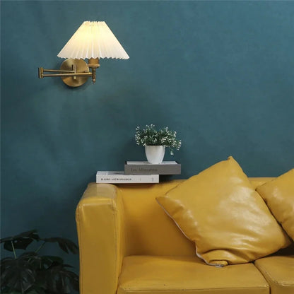 An adjustable brass swing arm wall lamp with a pleated white fabric shade, illuminated on a dark teal wall above a yellow leather sofa.