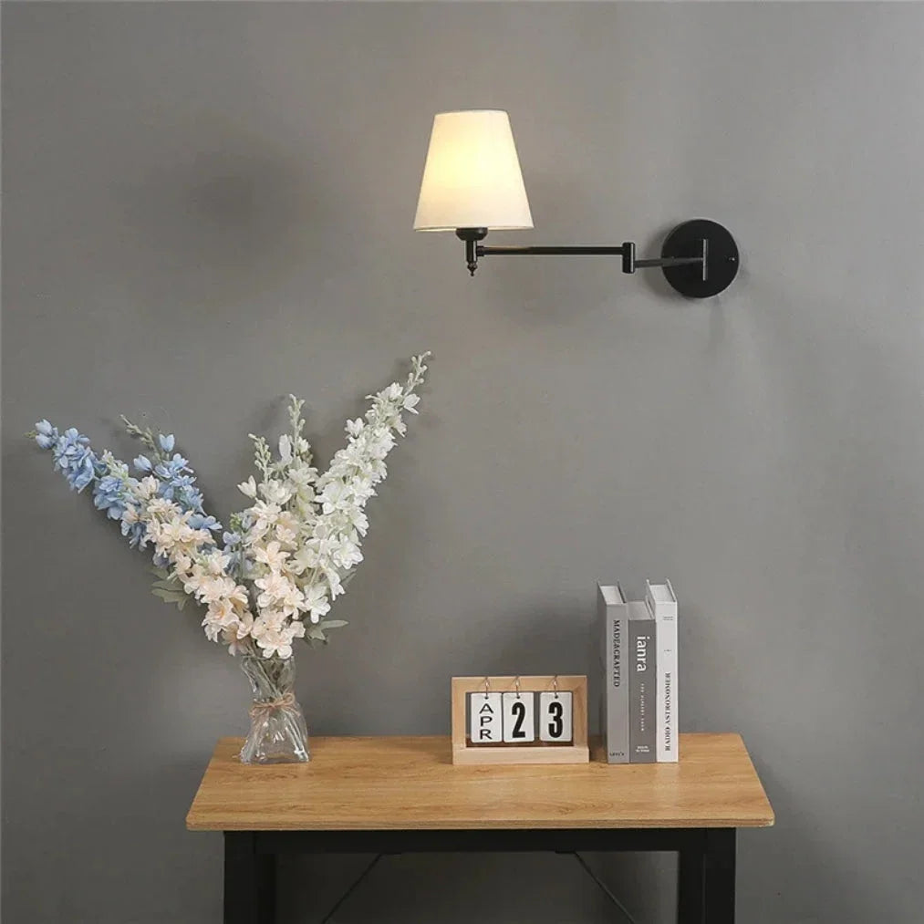A modern black swing arm wall lamp with a cream fabric shade, mounted on a grey wall above a wooden table with flowers and books.
