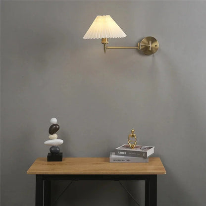 An adjustable brass swing arm wall lamp with a pleated white fabric shade, mounted on a grey wall above a wooden table with decorative objects.