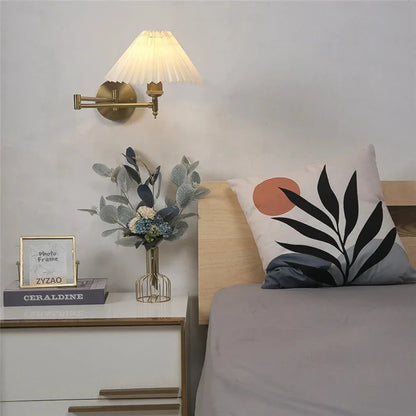 An adjustable brass swing arm wall lamp with a pleated white fabric shade, mounted above a bedside table as a cosy bedroom reading light.