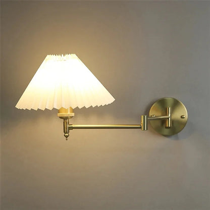 An adjustable brass swing arm wall lamp with a glowing white pleated fabric shade, mounted on a grey wall and casting a warm, soft light.