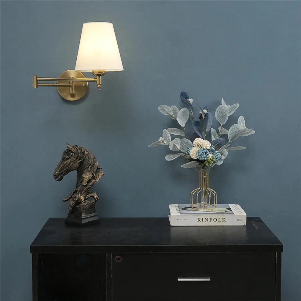 An adjustable brass swing arm wall lamp with a lit white fabric shade, mounted on a blue-grey wall above a black cabinet with home decor.