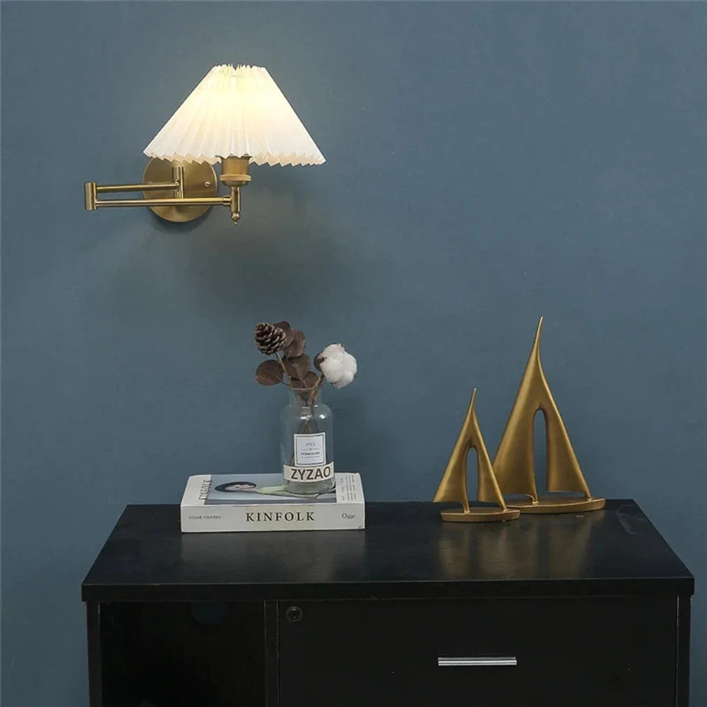 Brass adjustable swing arm wall lamp with a white pleated fabric shade, illuminated on a dark blue wall above a black cabinet with decorative items.