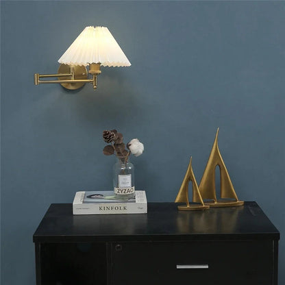 Brass adjustable swing arm wall lamp with a white pleated fabric shade, illuminated on a dark blue wall above a black cabinet with decorative items.