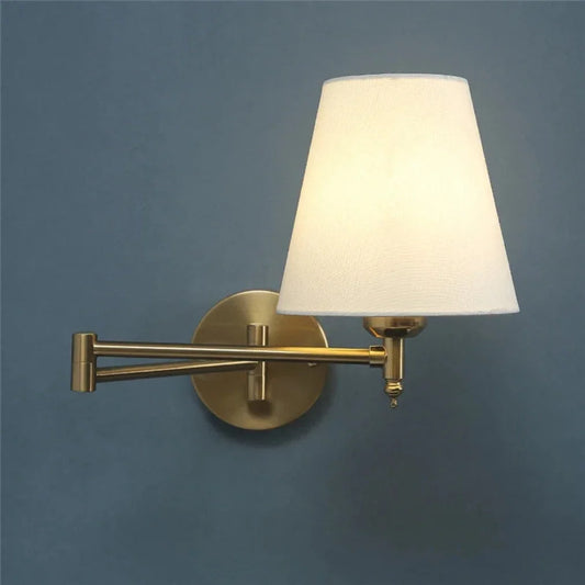 An adjustable swing arm wall lamp featuring a golden brass finish and a lit white fabric shade, mounted on a textured blue wall.
