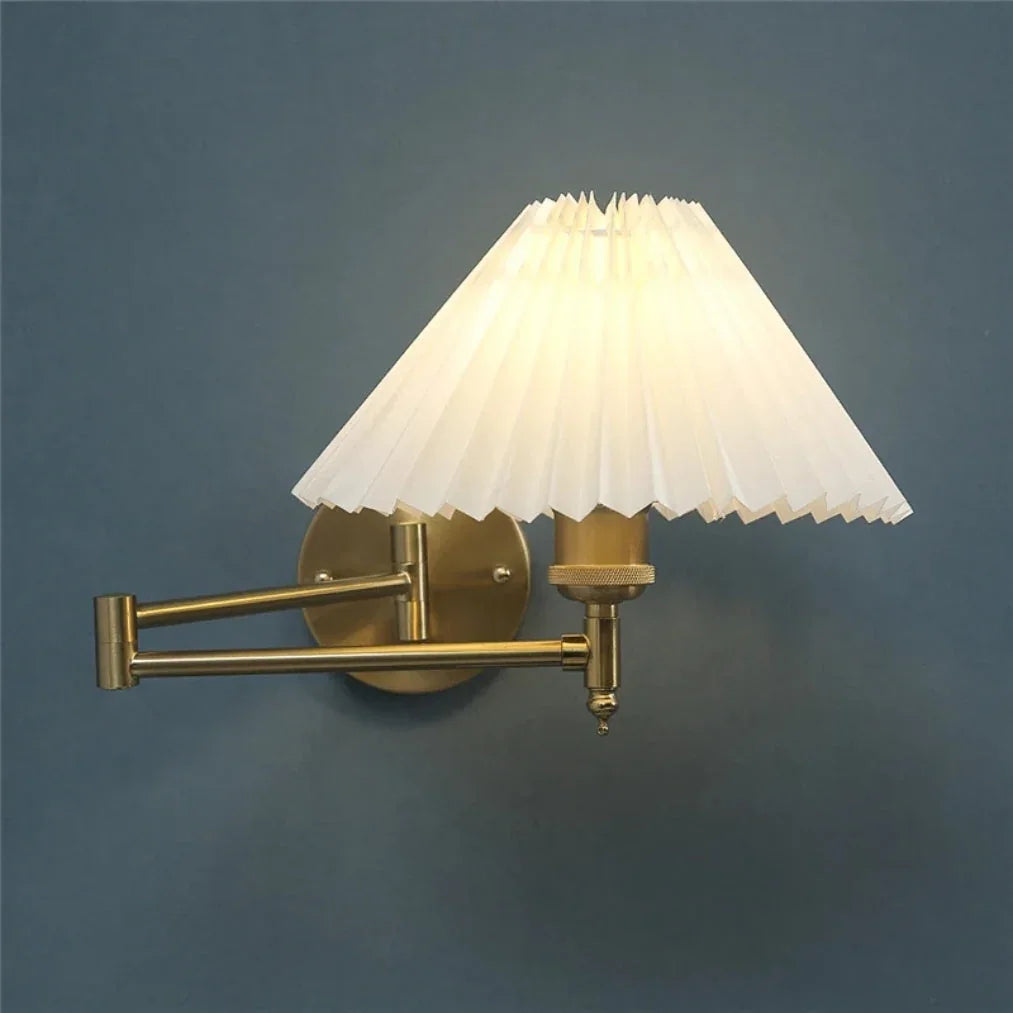An adjustable golden swing arm wall lamp with a glowing white pleated fabric shade, mounted on a dark blue wall to provide warm reading light.
