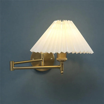 An adjustable golden swing arm wall lamp with a glowing white pleated fabric shade, mounted on a dark blue wall to provide warm reading light.