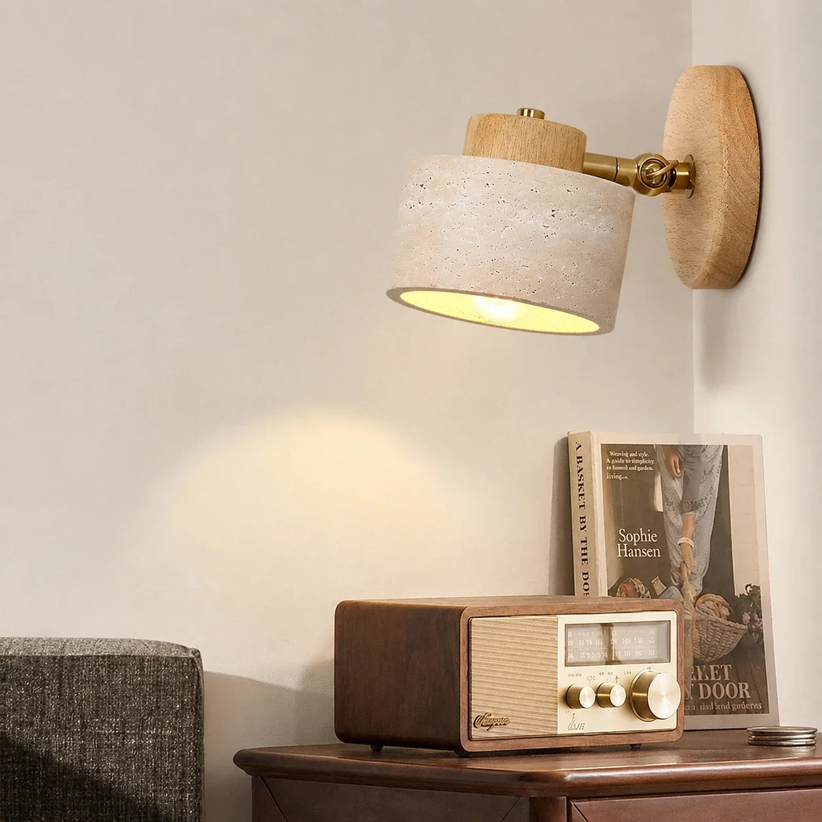 Adjustable wall light featuring a natural travertine shade and light wood mount, illuminating a bedside table with a vintage radio and a book.