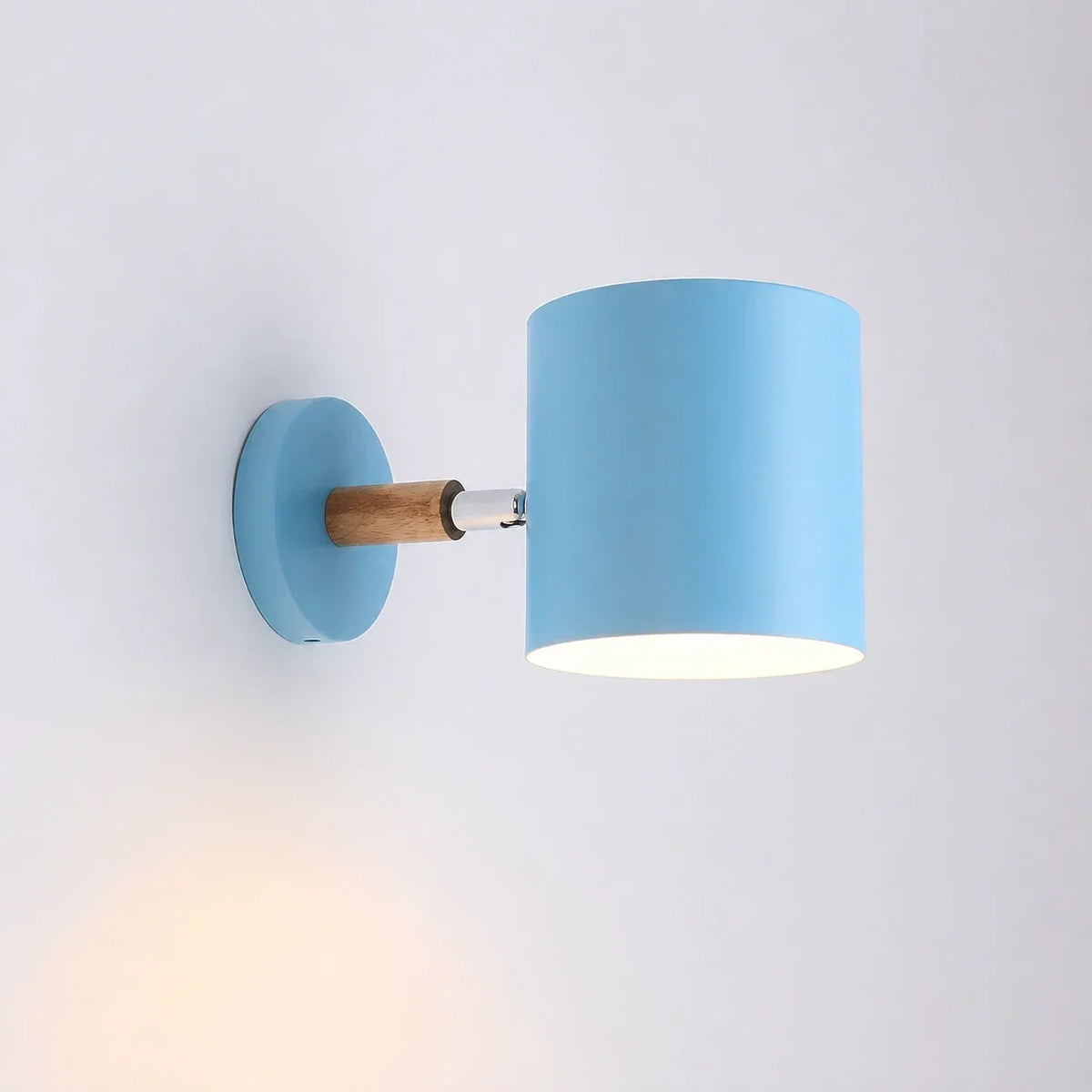 A modern light blue adjustable wall light with a cylindrical shade and a natural wood accent arm, mounted on a white wall and illuminated.