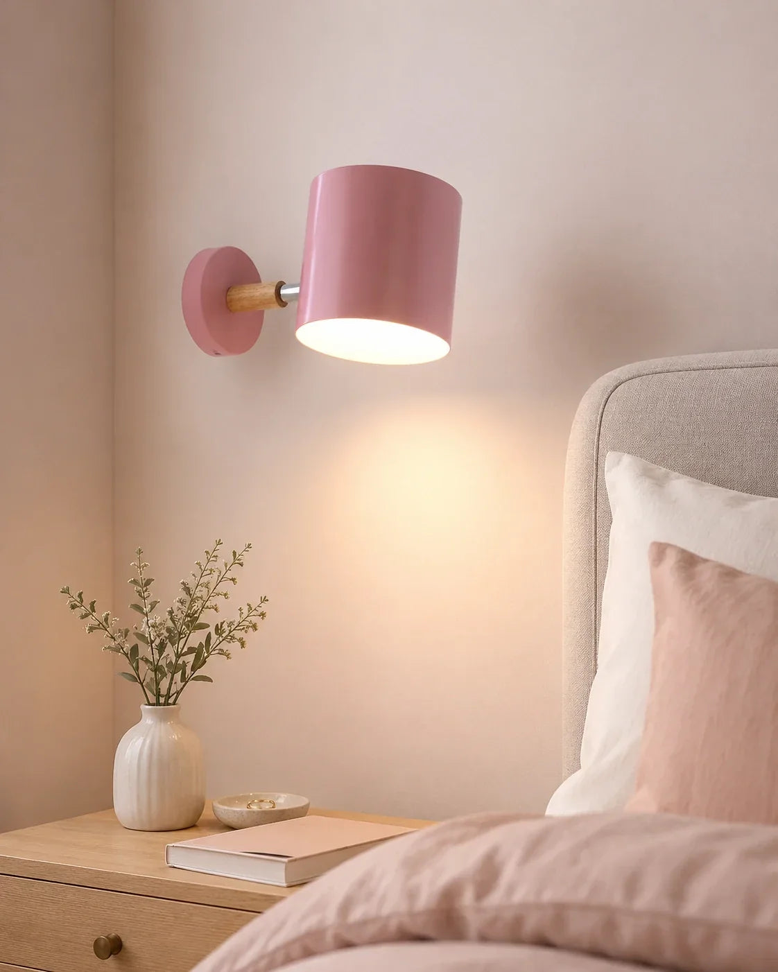 A pink adjustable wall light with a wood accent, mounted on a bedroom wall and casting a warm glow over a wooden bedside table.