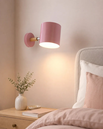 A pink adjustable wall light with a wood accent, mounted on a bedroom wall and casting a warm glow over a wooden bedside table.