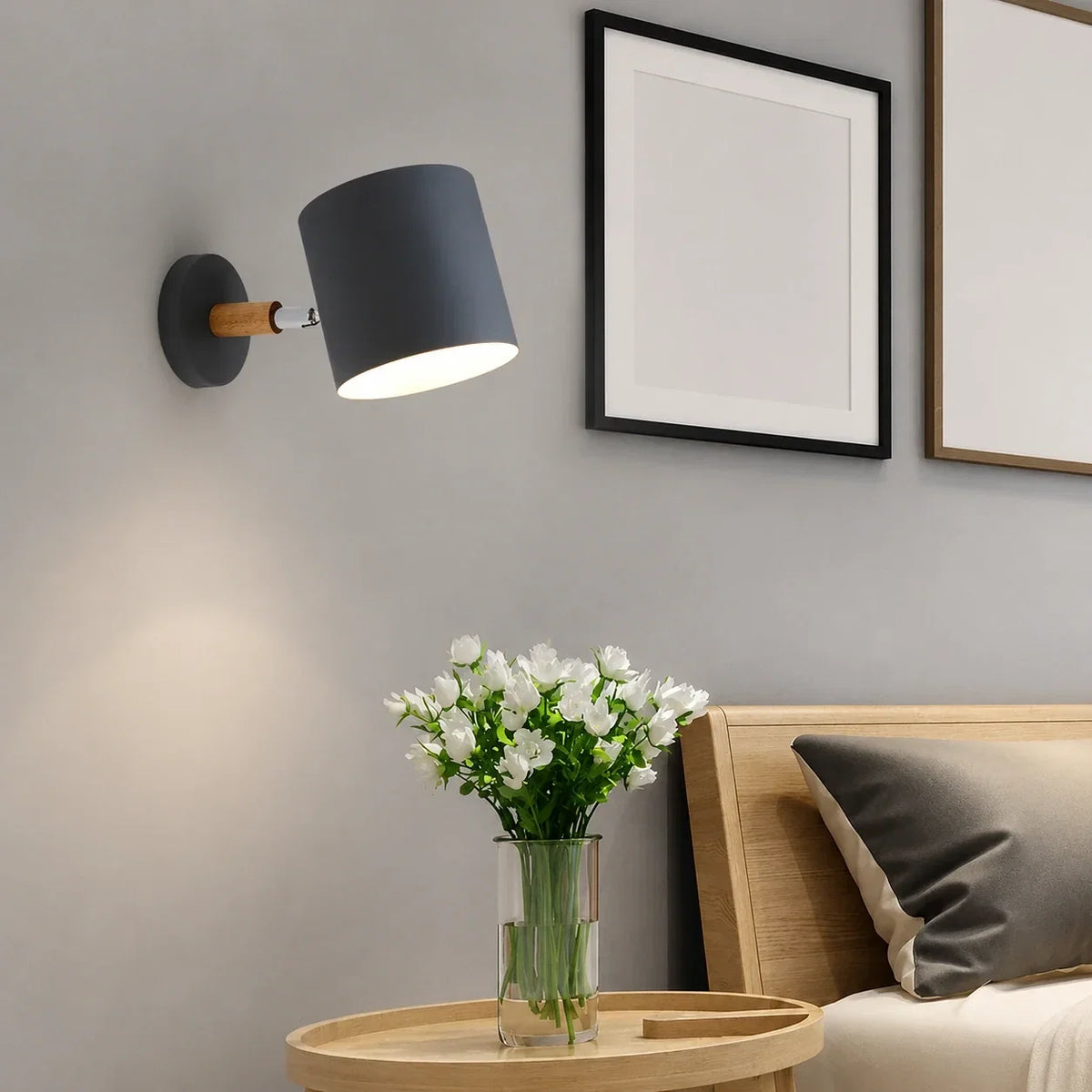 An adjustable grey wall light with a wood accent, illuminated above a wooden bedside table with white flowers in a modern bedroom setting.
