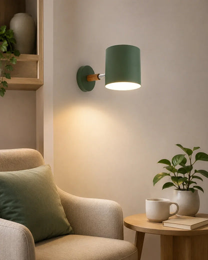 An adjustable green wall light with a wood accent, illuminated on a wall above a cosy armchair with a green cushion and a small wooden side table.