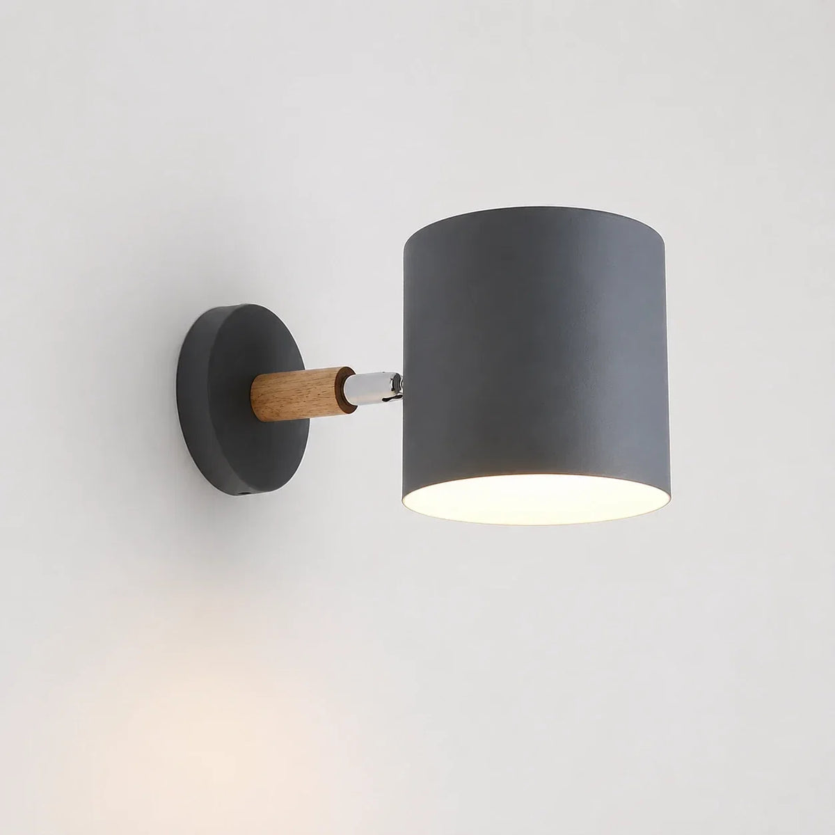Modern grey adjustable wall light with a cylindrical shade and a light wood accent, mounted on a white wall and illuminated with a warm glow.