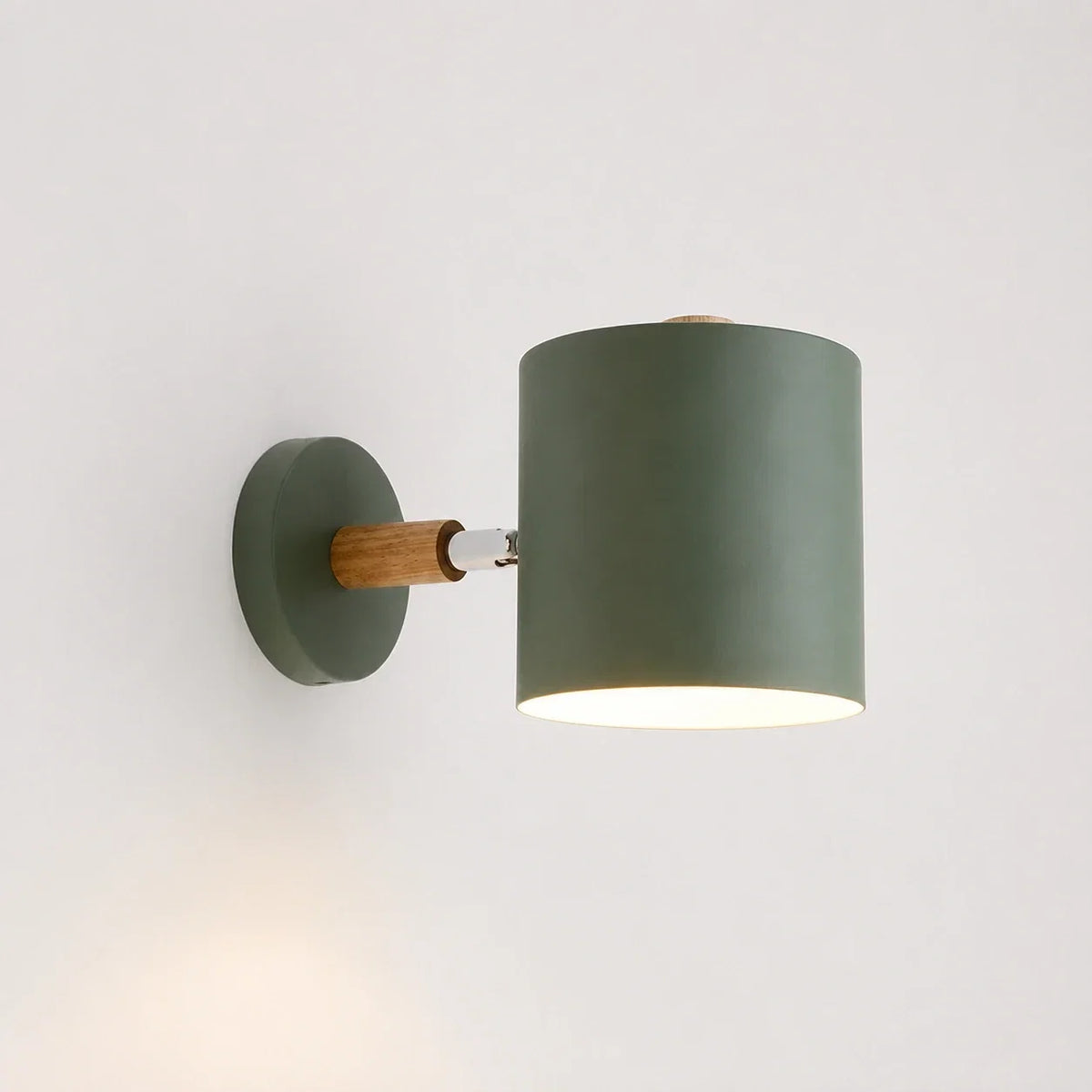 Modern green wall light with a cylindrical shade and a light wood accent, mounted on a white wall and illuminated with a warm downward glow.