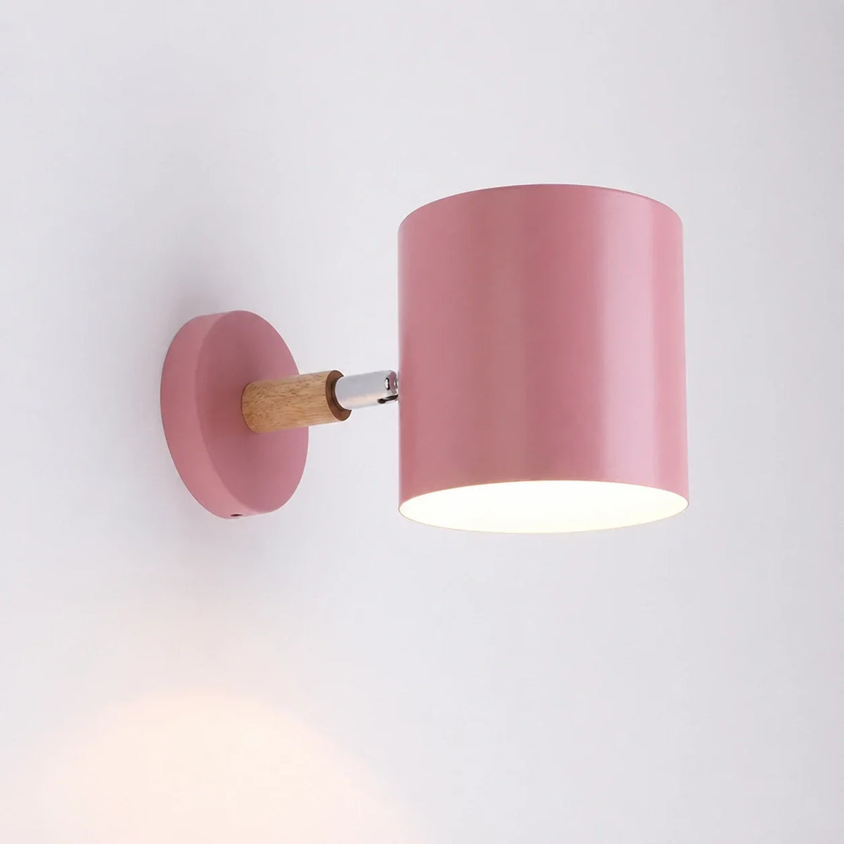A modern pink adjustable wall light with a cylindrical shade and a light wood accent, mounted on a white wall and illuminated.