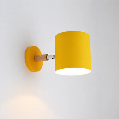 A modern yellow wall light with a cylindrical shade, a light wood accent, and a circular yellow base, illuminated and mounted on a plain white wall.