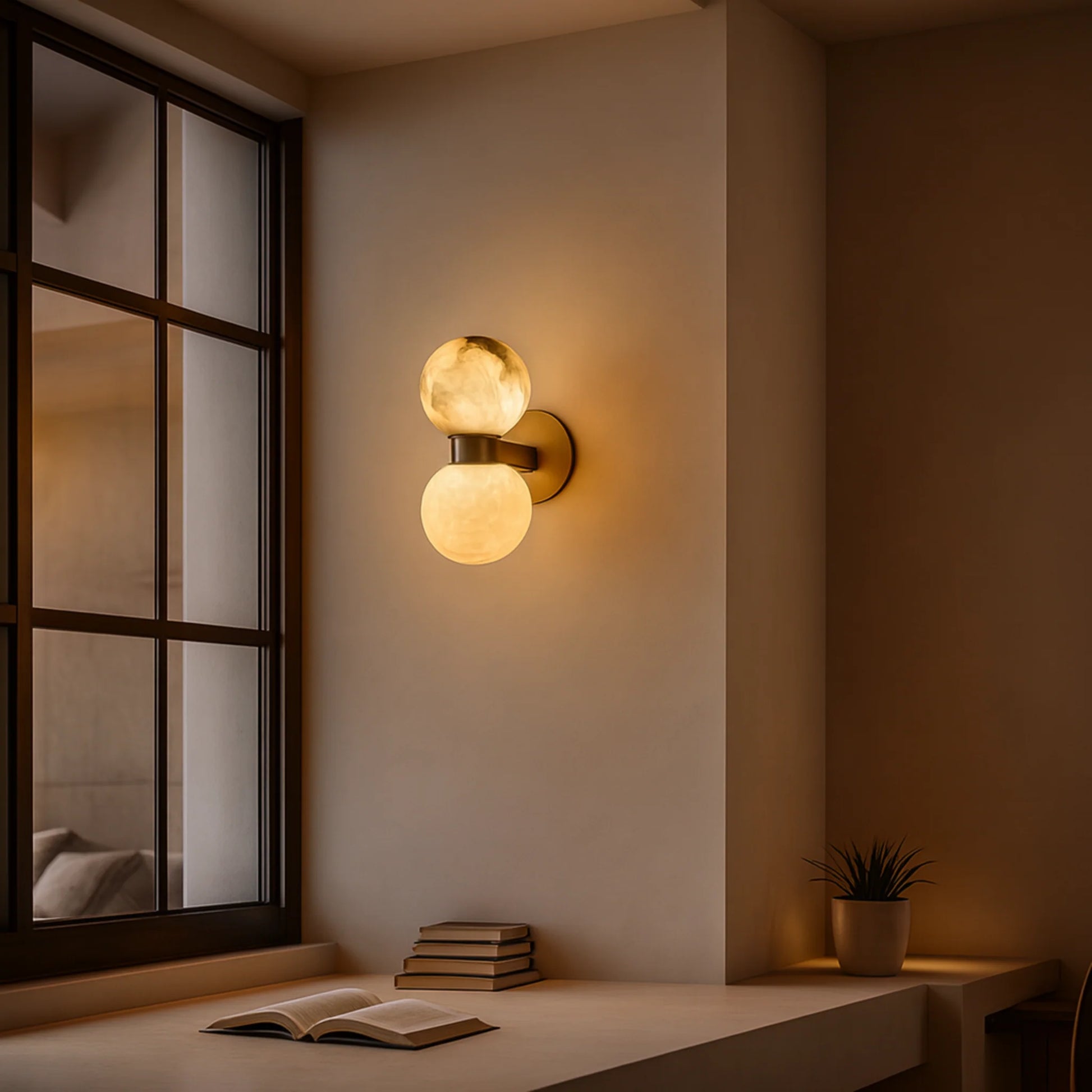Art Deco wall light featuring two glowing alabaster spheres mounted on a brushed brass base, illuminating a cosy interior space next to a window.