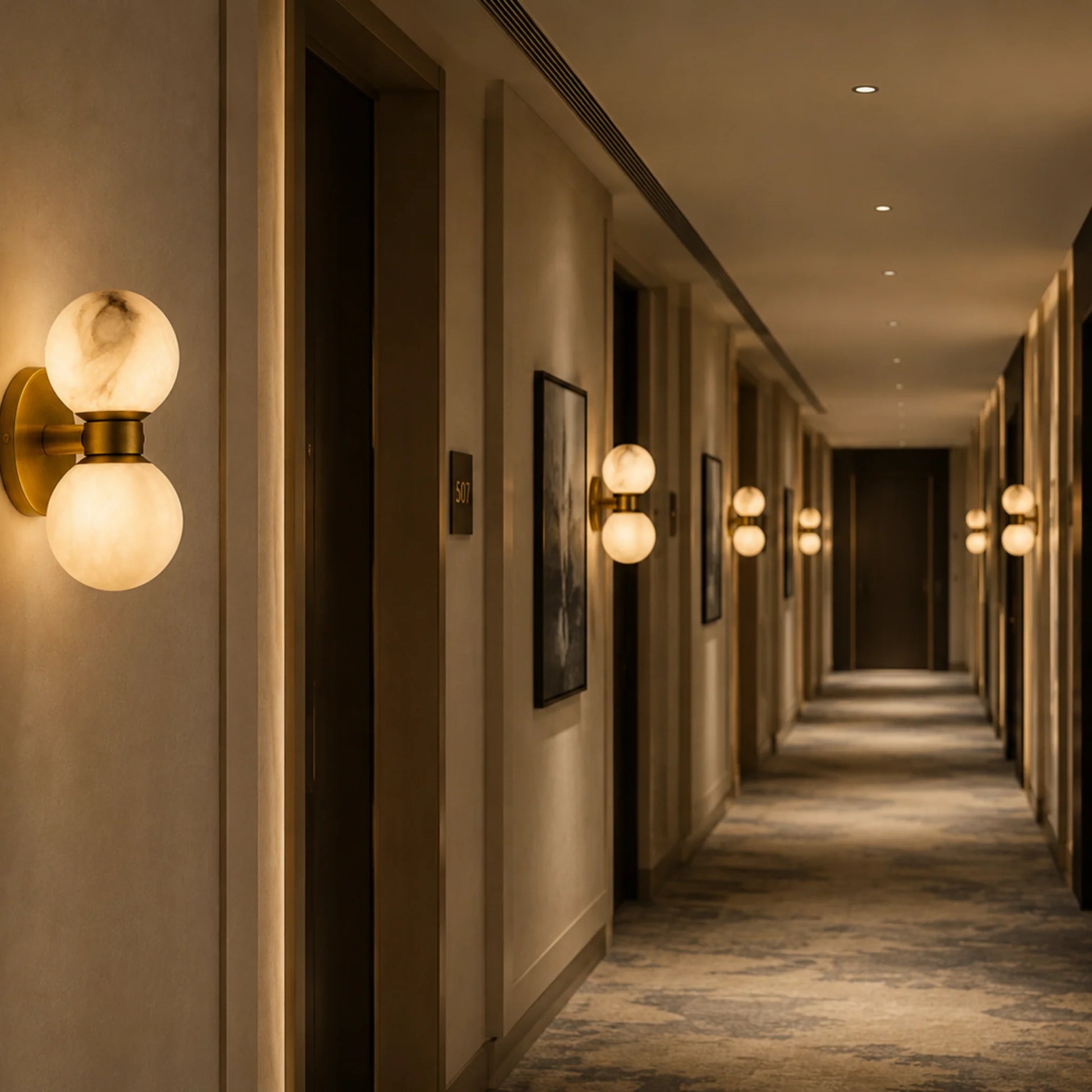 Art Deco wall lights with dual alabaster balls and brushed brass bases illuminating a long, elegant hotel corridor with warm ambient lighting.