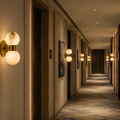 Art Deco wall lights with dual alabaster balls and brushed brass bases illuminating a long, elegant hotel corridor with warm ambient lighting.