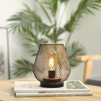 Battery operated LED table lamp featuring a decorative black metal mesh cage design, sitting on a stack of books with a blurred indoor plant background.