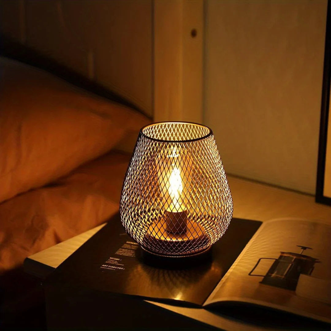 Battery operated LED table lamp with a decorative metal mesh design, casting a warm ambient glow on a bedside table next to an open book at night.