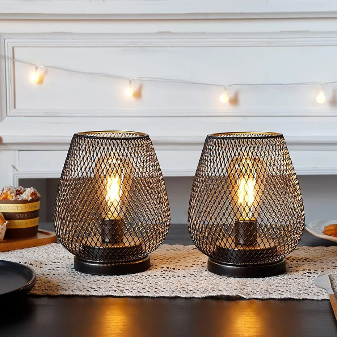 Two battery-operated LED table lamps with a black metal mesh design, glowing warmly on a dining table with a lace runner and fairy lights in the background