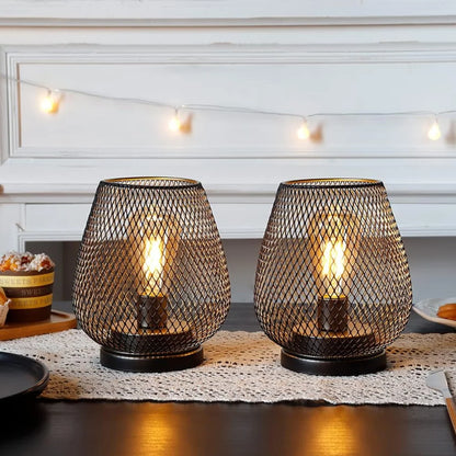 Two battery-operated LED table lamps with a black metal mesh design, glowing warmly on a dining table with a lace runner and fairy lights in the background