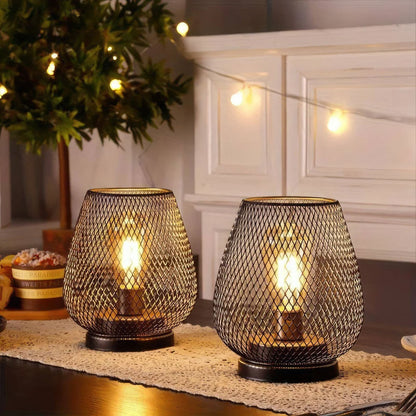 Two battery operated LED table lamps with a black wire mesh design, glowing warmly on a lace runner in a cosy, dimly lit room with ambient fairy lights.