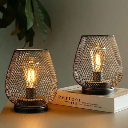 Two battery-operated LED table lamps with a dark metal mesh design and glowing filament bulbs, casting a warm ambient light on a wooden surface.