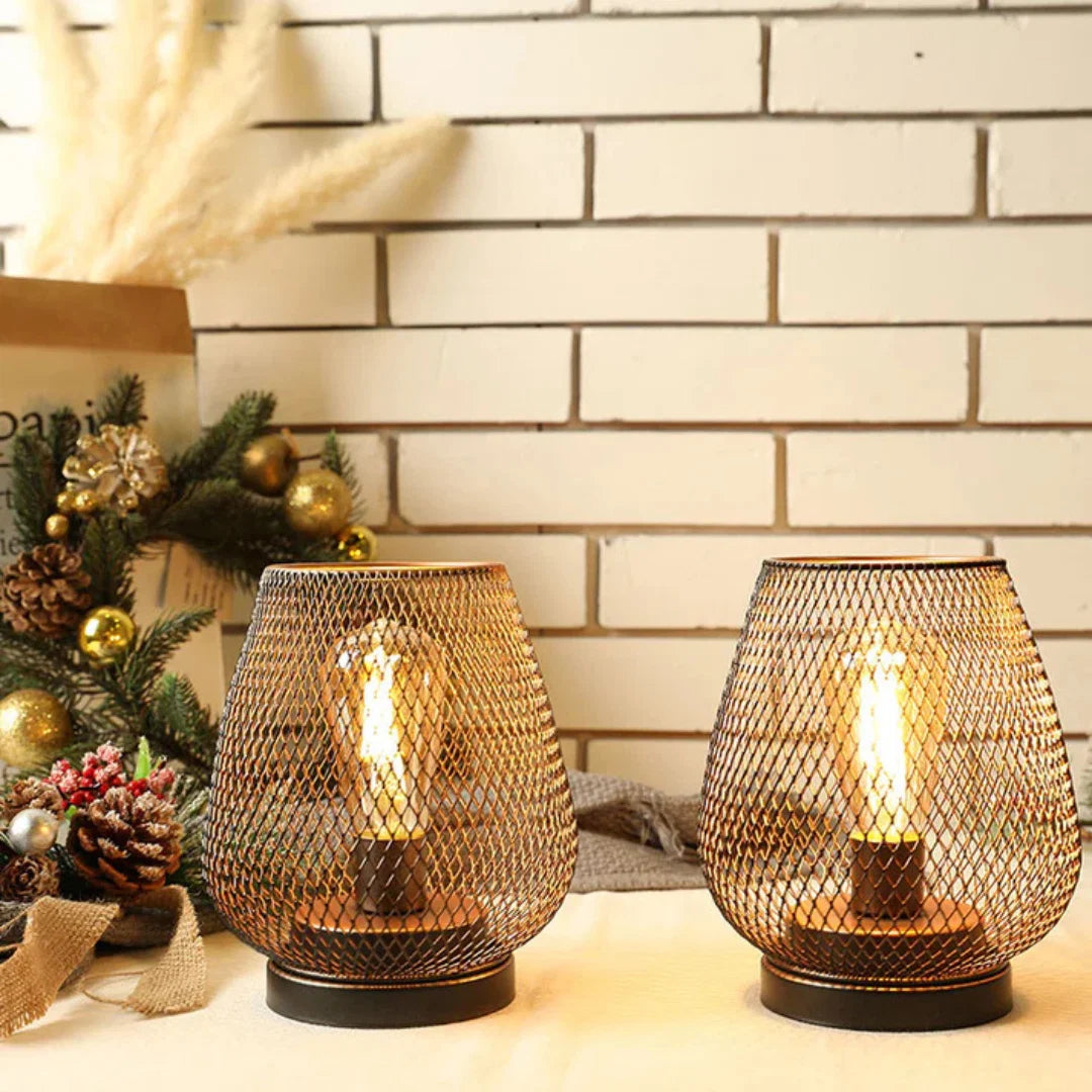 Two battery-operated LED table lamps with a decorative metal mesh design, glowing warmly on a table against a white brick wall with festive decor.