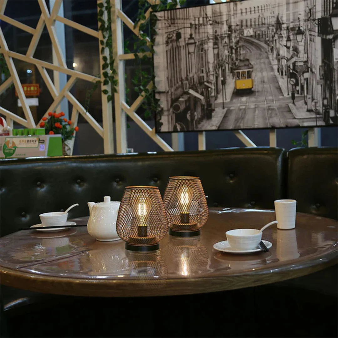 Two battery-operated LED table lamps with a decorative bronze mesh design glowing on a round dining table in a cosy restaurant setting.