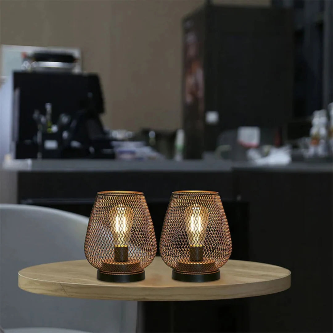 Two battery-operated LED table lamps with a decorative bronze mesh design, glowing warmly on a round wooden table in a contemporary interior setting.