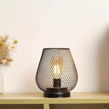 Battery operated table lamp with a black metal mesh shade and glowing LED bulb, creating ambient lighting on a wooden shelf with dried flowers nearby.