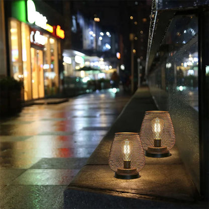 Two battery-operated LED table lamps with a decorative metal mesh design glowing on a stone ledge at night with a blurred city street in the background.