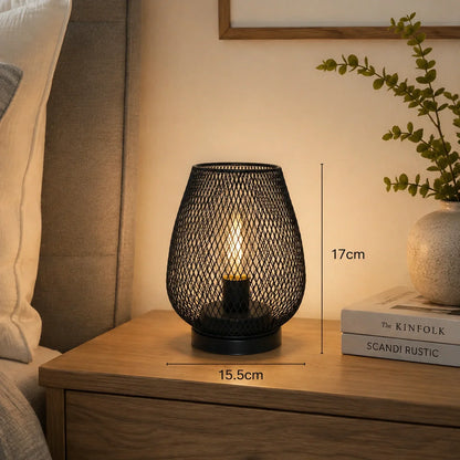 Black battery-operated LED table lamp with a decorative mesh design, measuring 17cm in height and 15.5cm in width, displayed on a wooden bedside table.