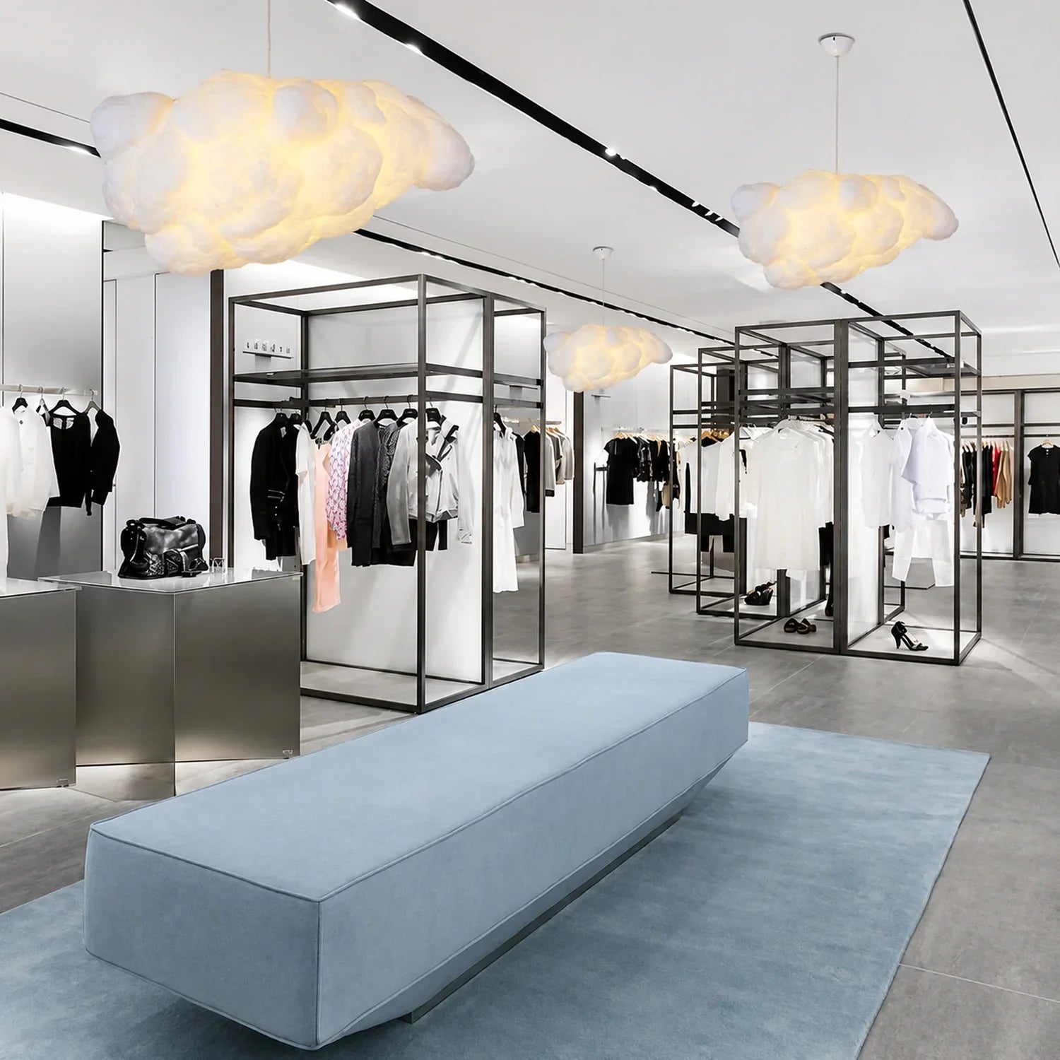 Three sculptural cloud pendant lights glowing in a modern clothing boutique with minimalist black clothing rails and a light blue bench on a rug.