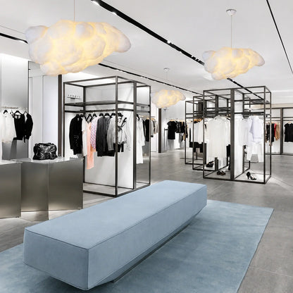 Three sculptural cloud pendant lights glowing in a modern clothing boutique with minimalist black clothing rails and a light blue bench on a rug.