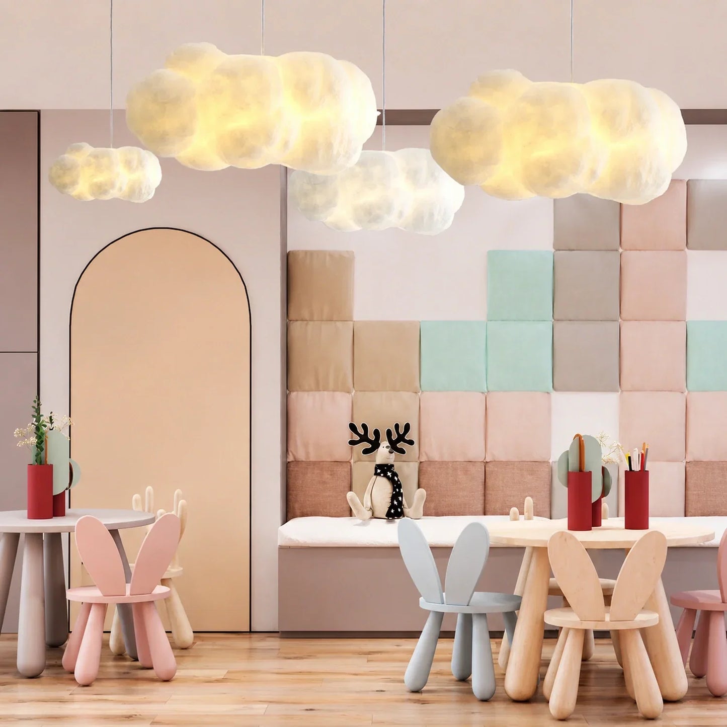 Four cloud-shaped pendant lights hanging in a modern children's playroom with pastel-coloured wall panels, wooden tables and bunny-ear chairs.