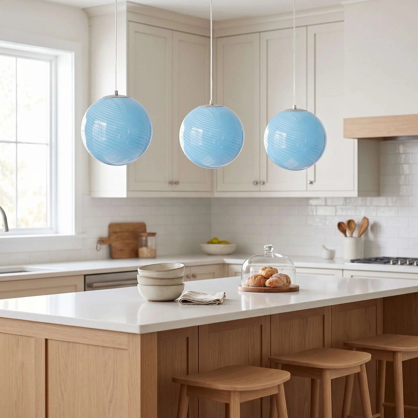 Three modern light blue swirl glass pendant lights hanging in a row above a kitchen island with wooden stools and a bright, contemporary interior.