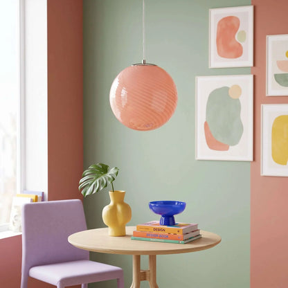A round, pink swirl glass pendant light hangs above a small wooden dining table in a colourful room with abstract wall art and a lilac chair.