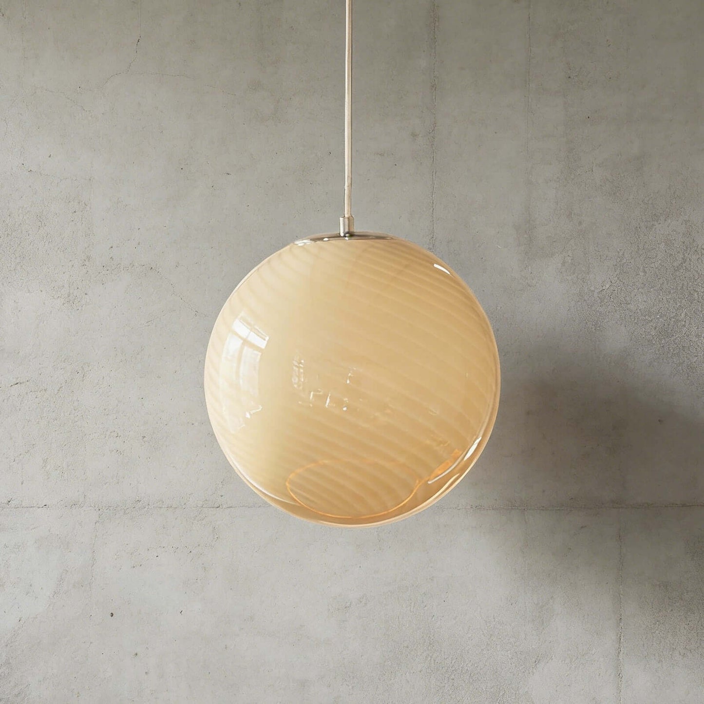A modern spherical pendant light with a subtle cream swirl glass shade, suspended by a simple metal rod against a textured grey concrete wall.