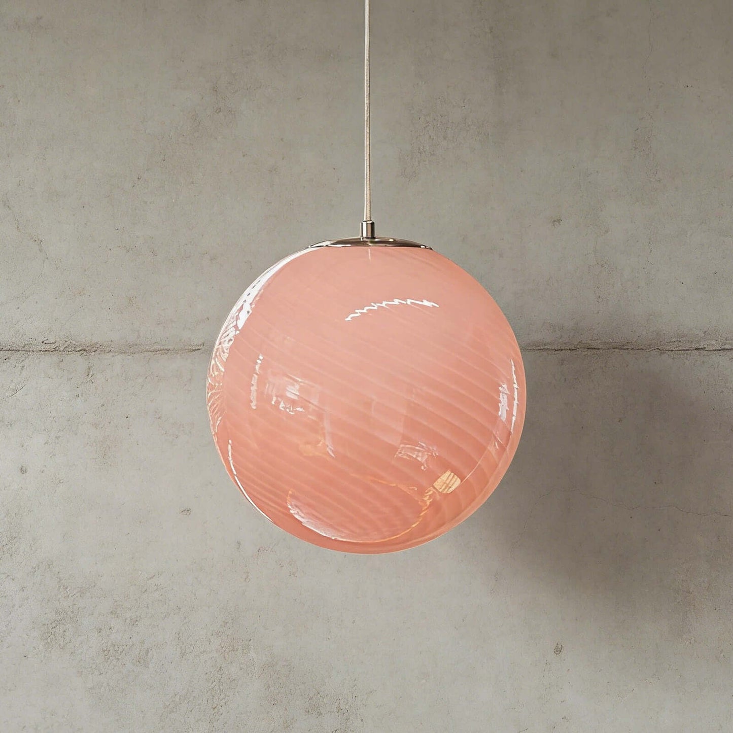 A modern spherical pendant light featuring a soft pink glass shade with a subtle swirl pattern, suspended against a neutral grey concrete wall.