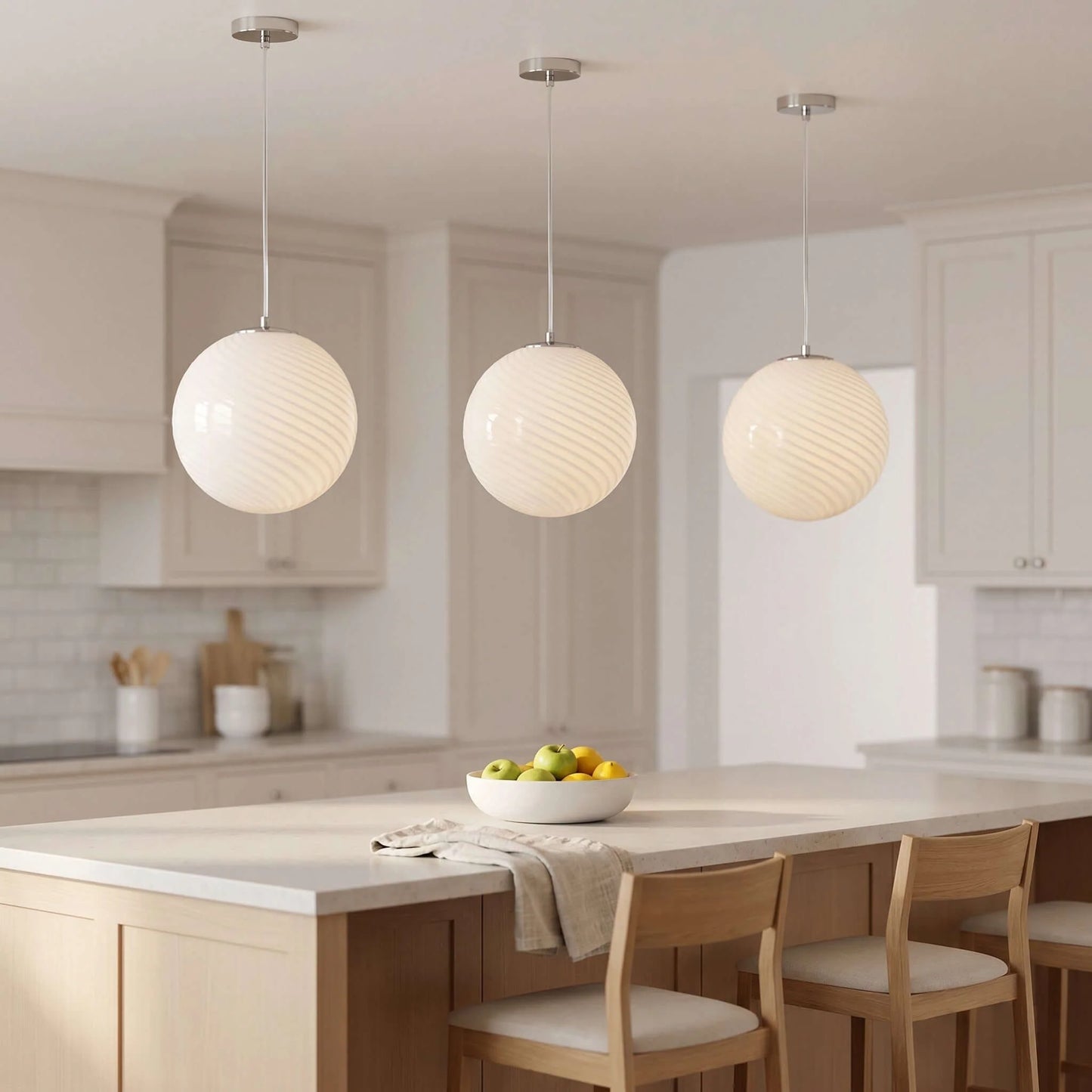 Three white swirl glass pendant lights hanging above a kitchen island with wooden chairs and a bowl of fruit in a modern, neutral-toned kitchen.