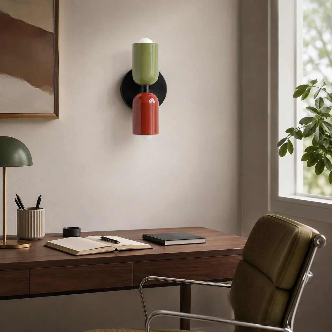 Contemporary dual-cylinder wall lamp with green and red shades mounted above a wooden desk in a modern home office setting with natural light.