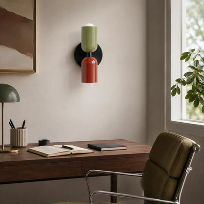 Contemporary dual-cylinder wall lamp with green and red shades mounted above a wooden desk in a modern home office setting with natural light.